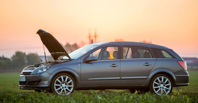 Station wagon with the hood up parked on the side of the road with the city in the background